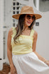 Woman wearing a yellow top and white skirt with a straw hat and sunglasses, standing against a blurred outdoor background.